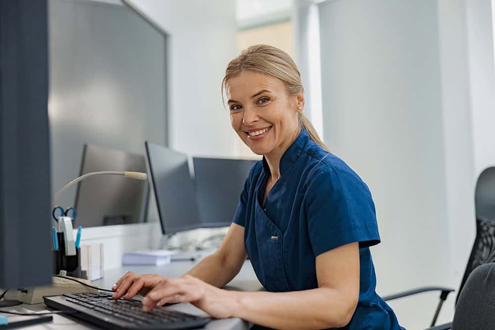 A Woman In Blue Scrubs Is Smiling While Working On A Computer.